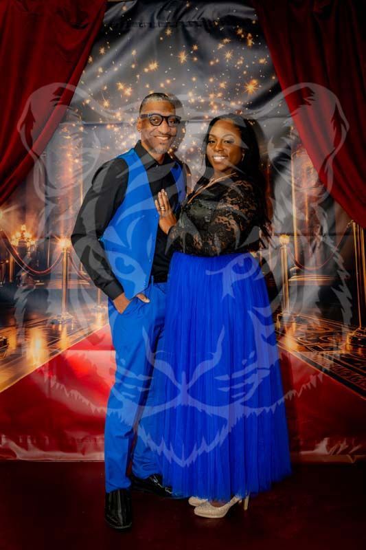 A couple posing in blue formal wear against a red-curtained, gala-themed backdrop with a faint tiger overlay.