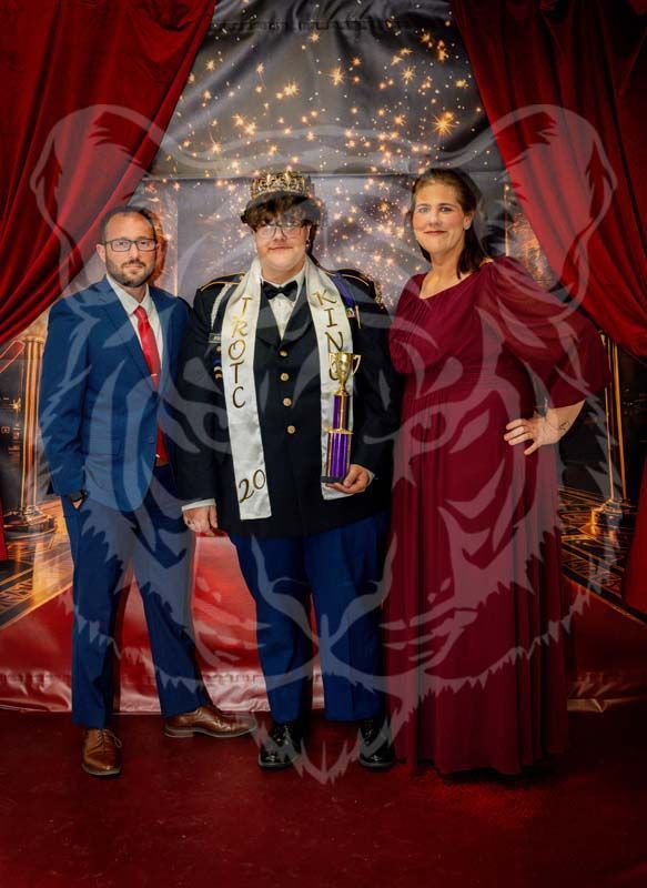 Three people pose for a photo in front of a red curtain, with the center person wearing a crown, sash, and holding a trophy.