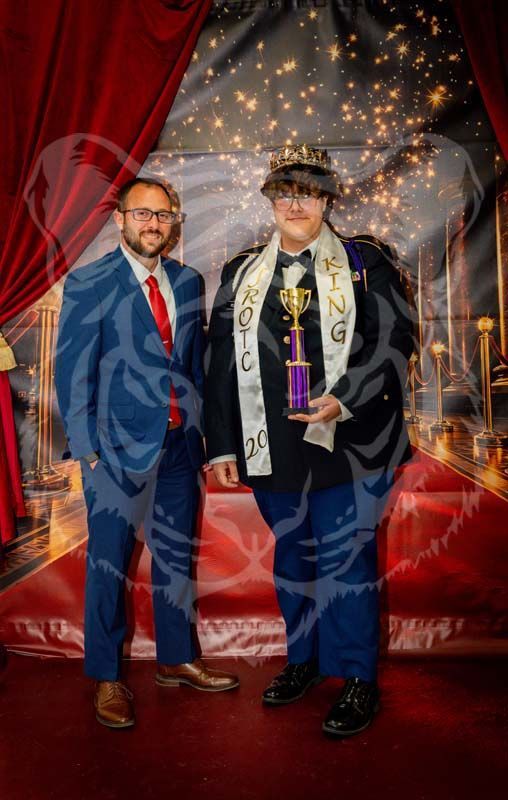Two individuals in formal attire pose in front of a red-curtained stage. One wears a crown and sash while holding a trophy.