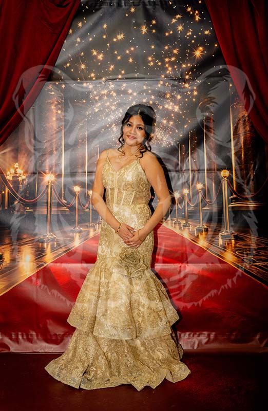 A person in a gold, floor-length mermaid gown stands before a backdrop of a red carpet, curtains, and twinkling lights.