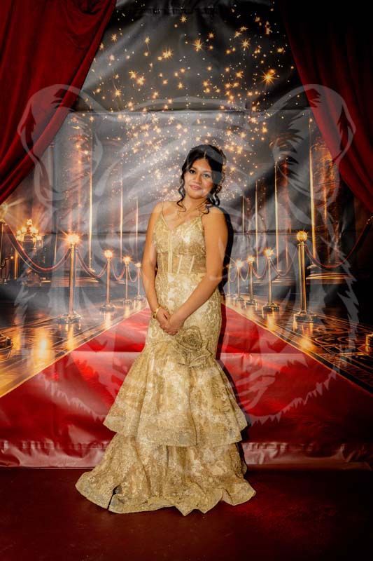 A person in a gold, floor-length dress poses in front of a red-carpet-themed backdrop with golden lights.