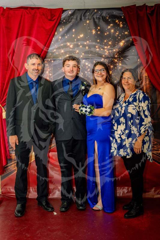 Four people stand posed for a photo in front of a red-curtained, string-lit backdrop; two men in suits, a woman in a gown.