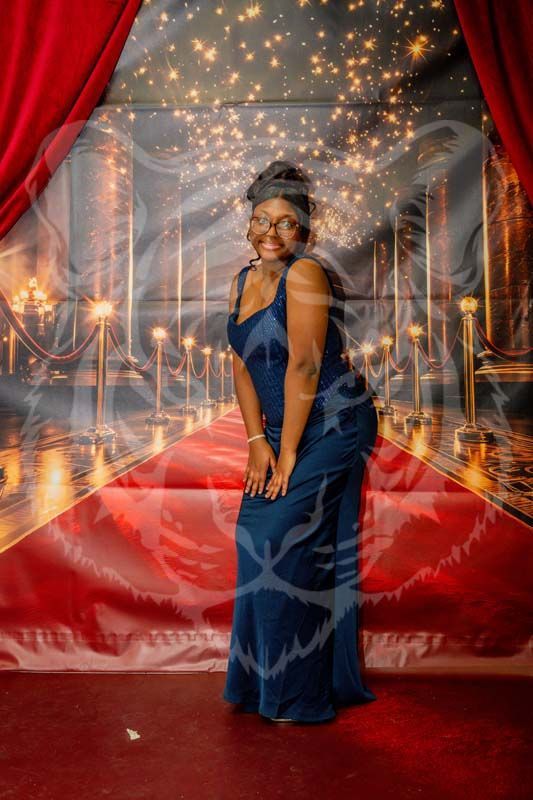 A person in a long dark blue dress smiling on a red carpet backdrop with sparkling lights and velvet curtains.