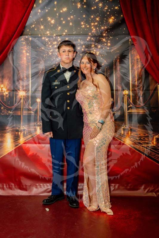 A young person in a dark formal military uniform stands next to a person in a sequined, floor-length champagne gown.
