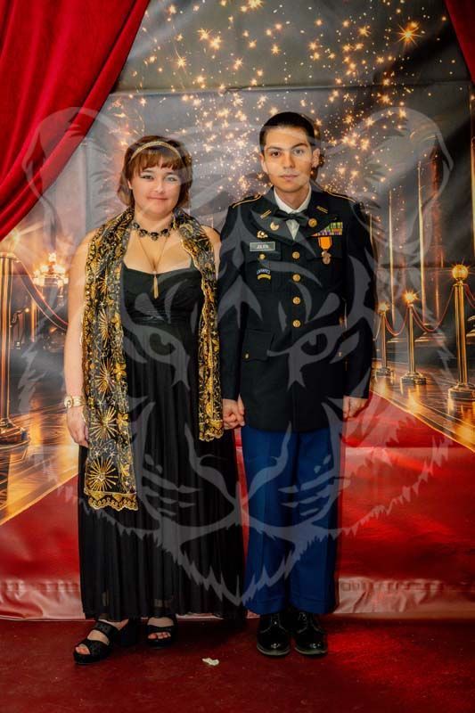 A person in a long black dress and patterned scarf stands beside a soldier in a dress uniform before a red carpet backdrop.