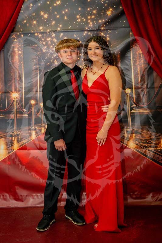 A couple stands together for a formal photo in front of a red-curtained backdrop with fairy lights.