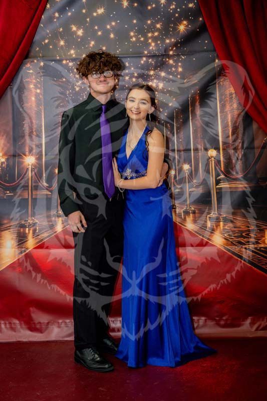 A person in a black suit and purple tie stands with a person in a blue formal gown before a red-curtained backdrop.