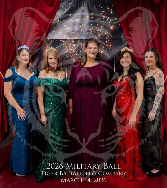 Five people in evening gowns and tiaras pose in front of a red-curtained backdrop with a tiger graphic for a military ball.
