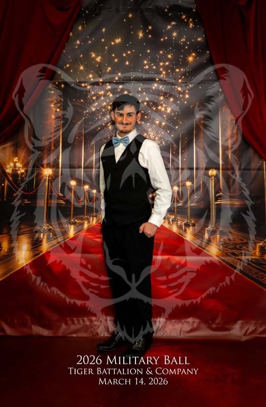 A person wearing a suit and bowtie stands on a red carpet in front of a 2026 Military Ball banner with a tiger watermark.