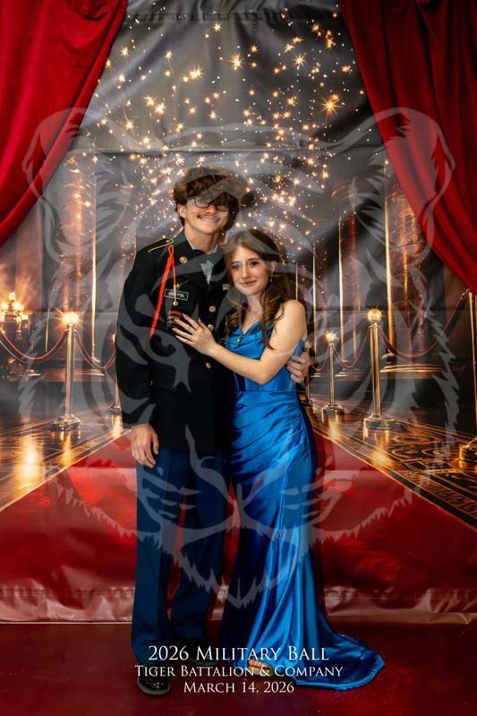 A couple poses together in formal wear before a backdrop of red curtains, lights, and a red carpet for a military ball.