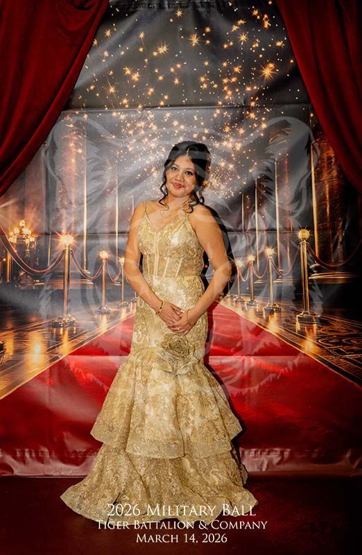 A person in a floor-length gold gown posing in front of a red carpet and red curtain backdrop for a 2026 military ball.