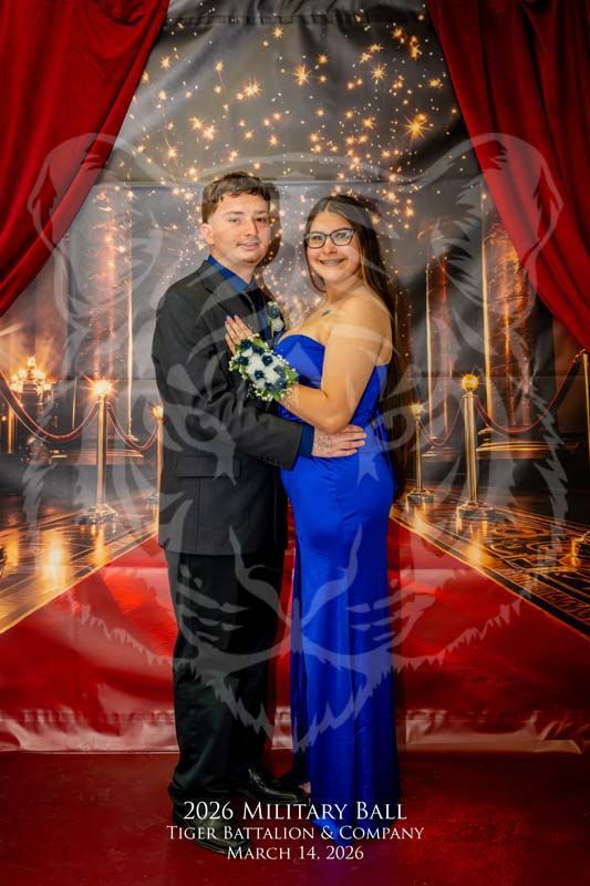 A couple poses for a photo at the 2026 Military Ball in front of a red curtain and ballroom backdrop.