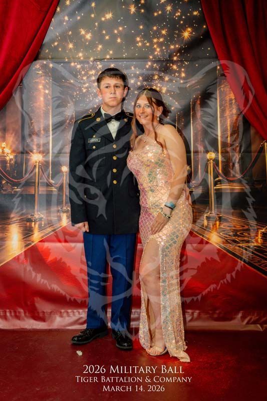 A couple in formal attire stands on a red carpet in front of a backdrop with lights, text, and a stylized tiger overlay.