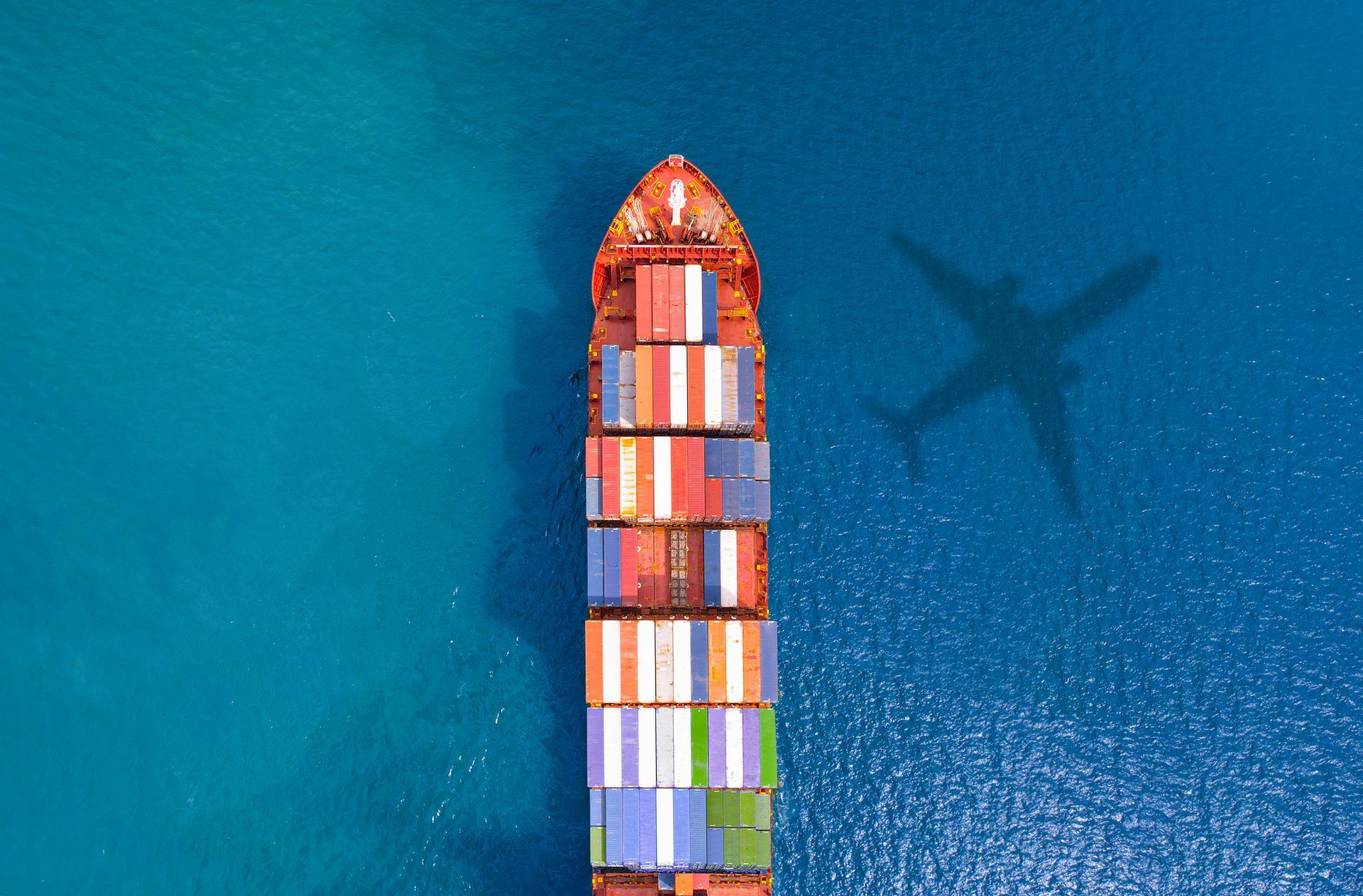 An aerial view of a large container ship in the ocean with a plane shadow on the water.