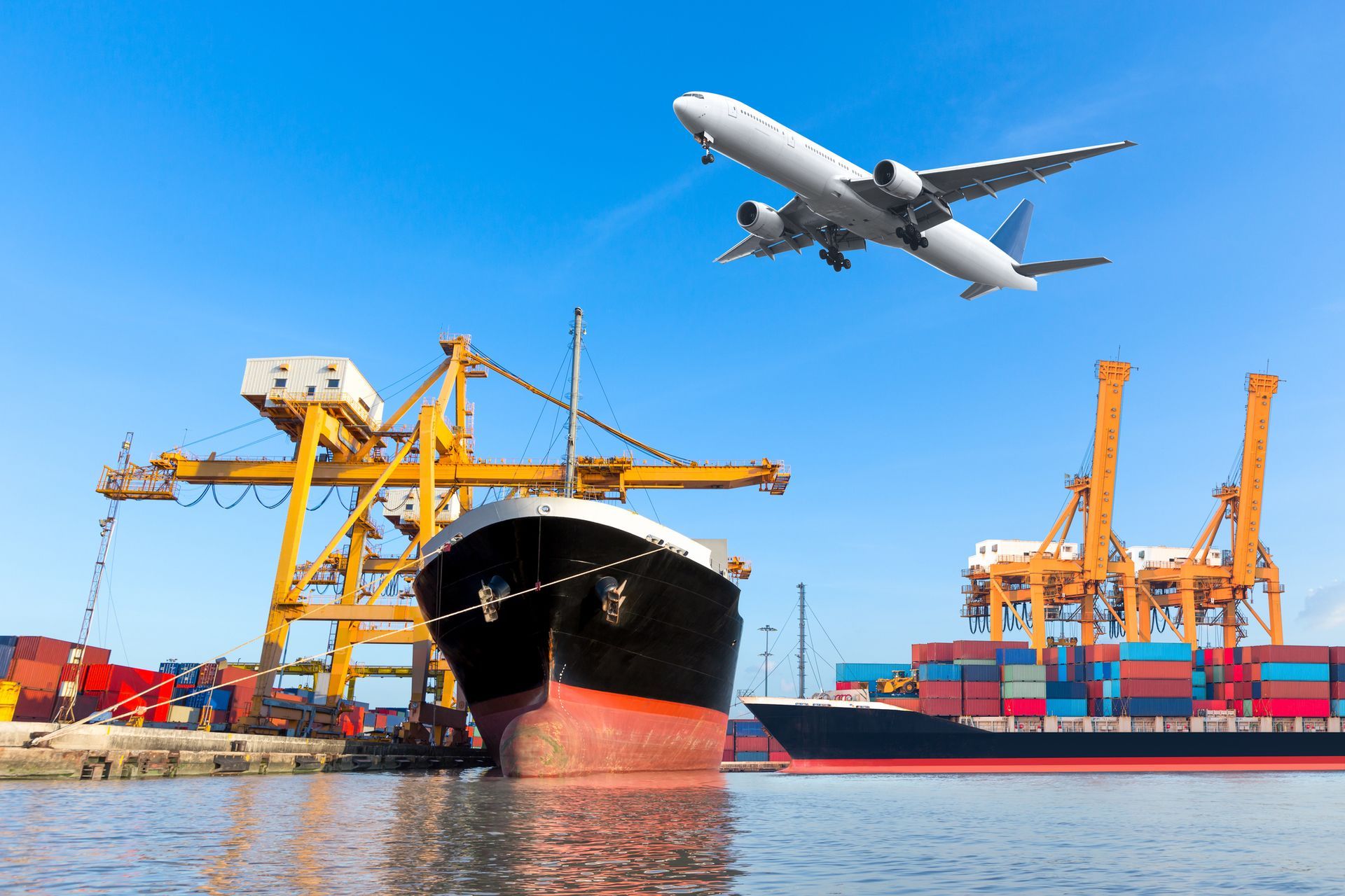 An airplane is flying over a cargo ship in a harbor.