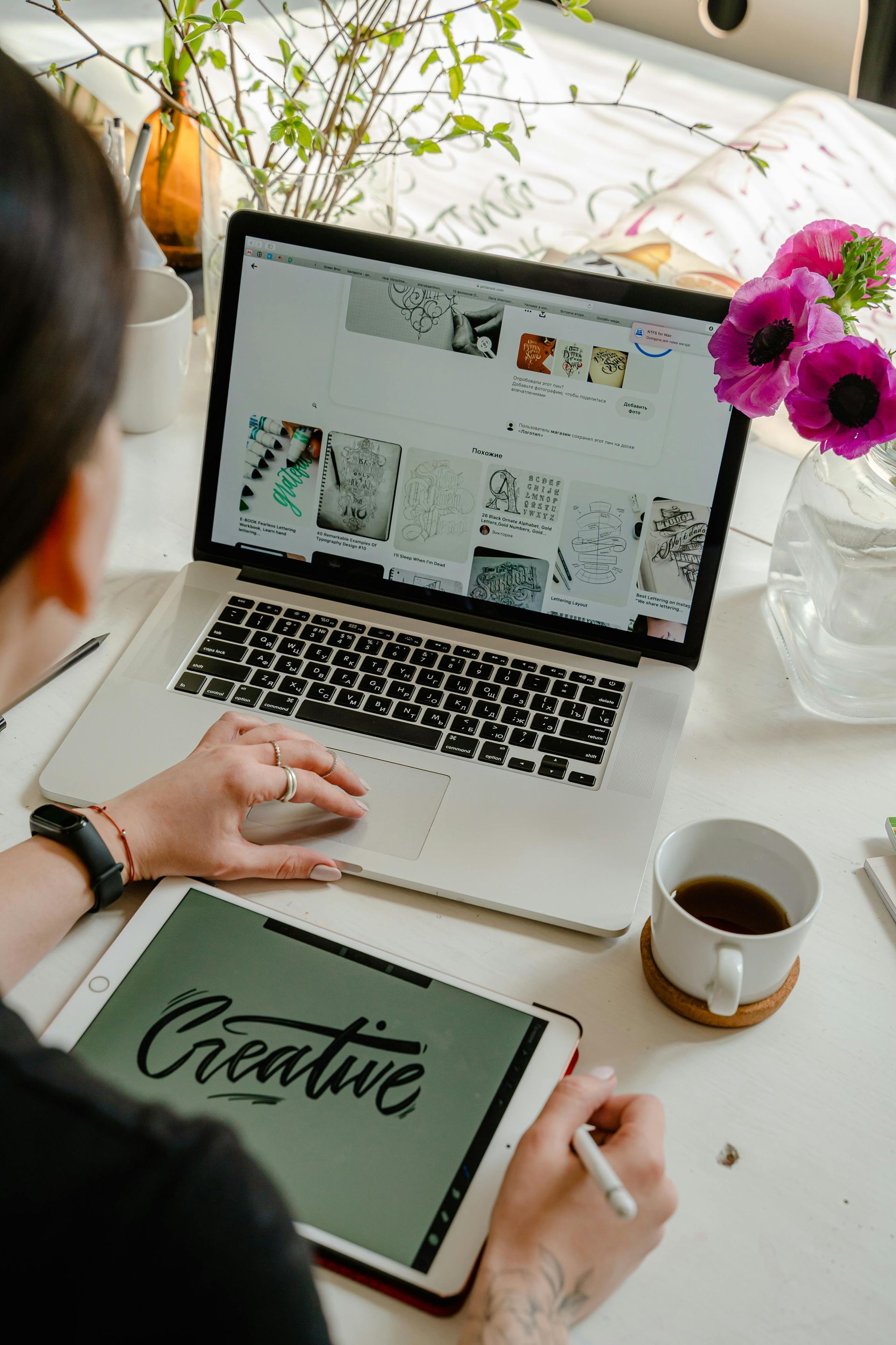 Person working at a desk with a laptop and tablet, flowers, and a coffee cup.