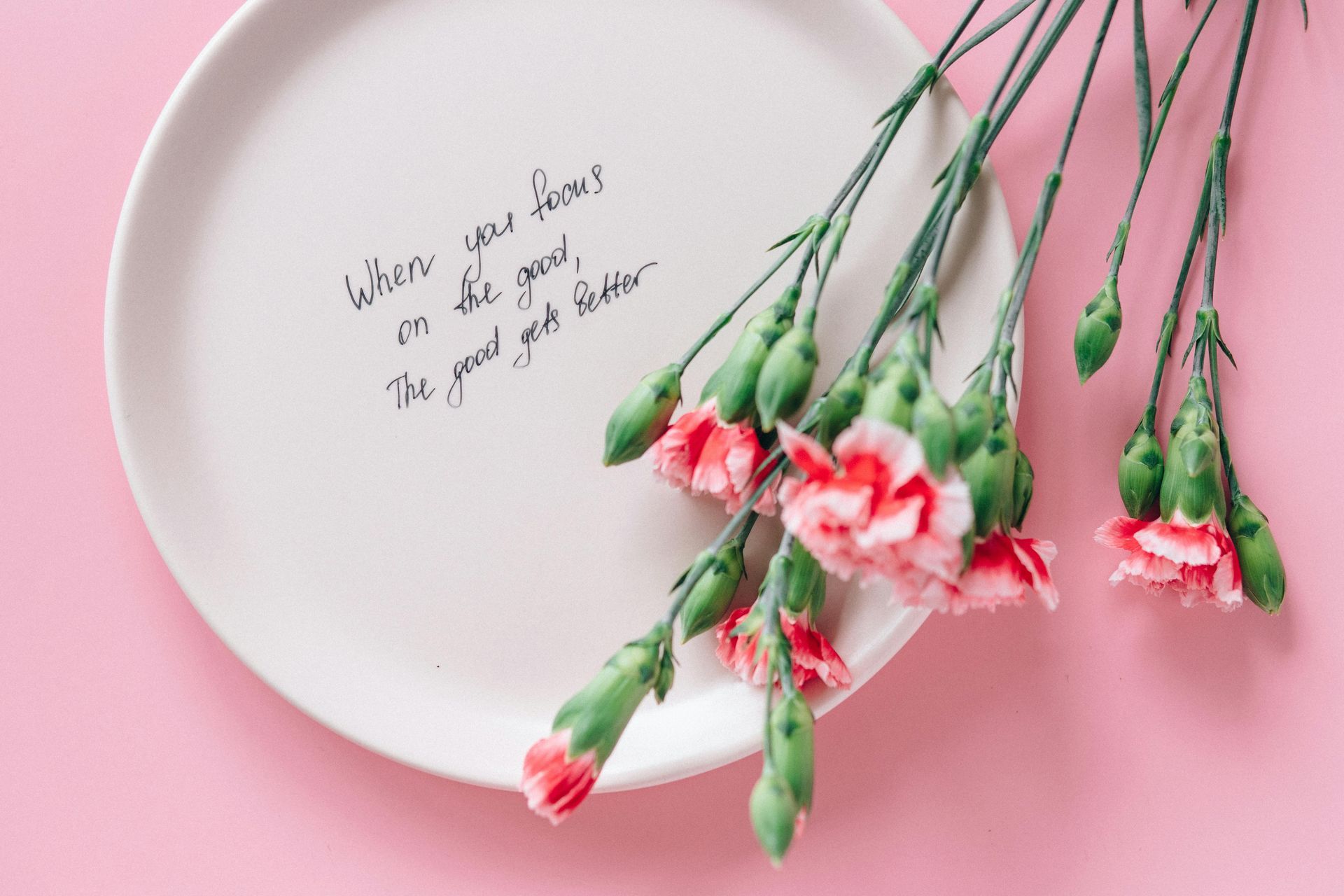 White plate with handwritten text and pink flowers on a pink background.