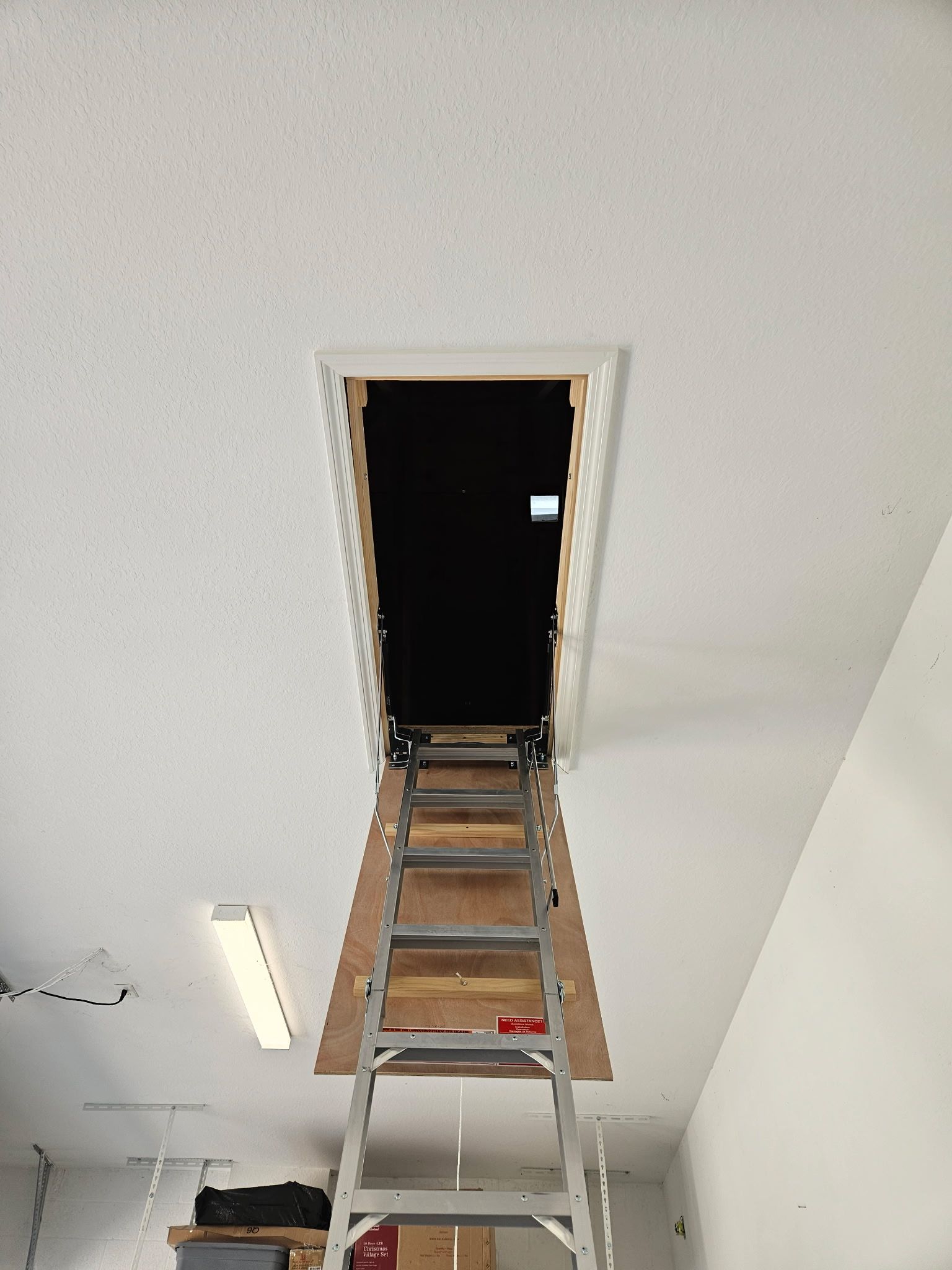 A folding attic ladder extends from a ceiling opening. The ladder is wood-toned and steel. The opening leads to a dark space.