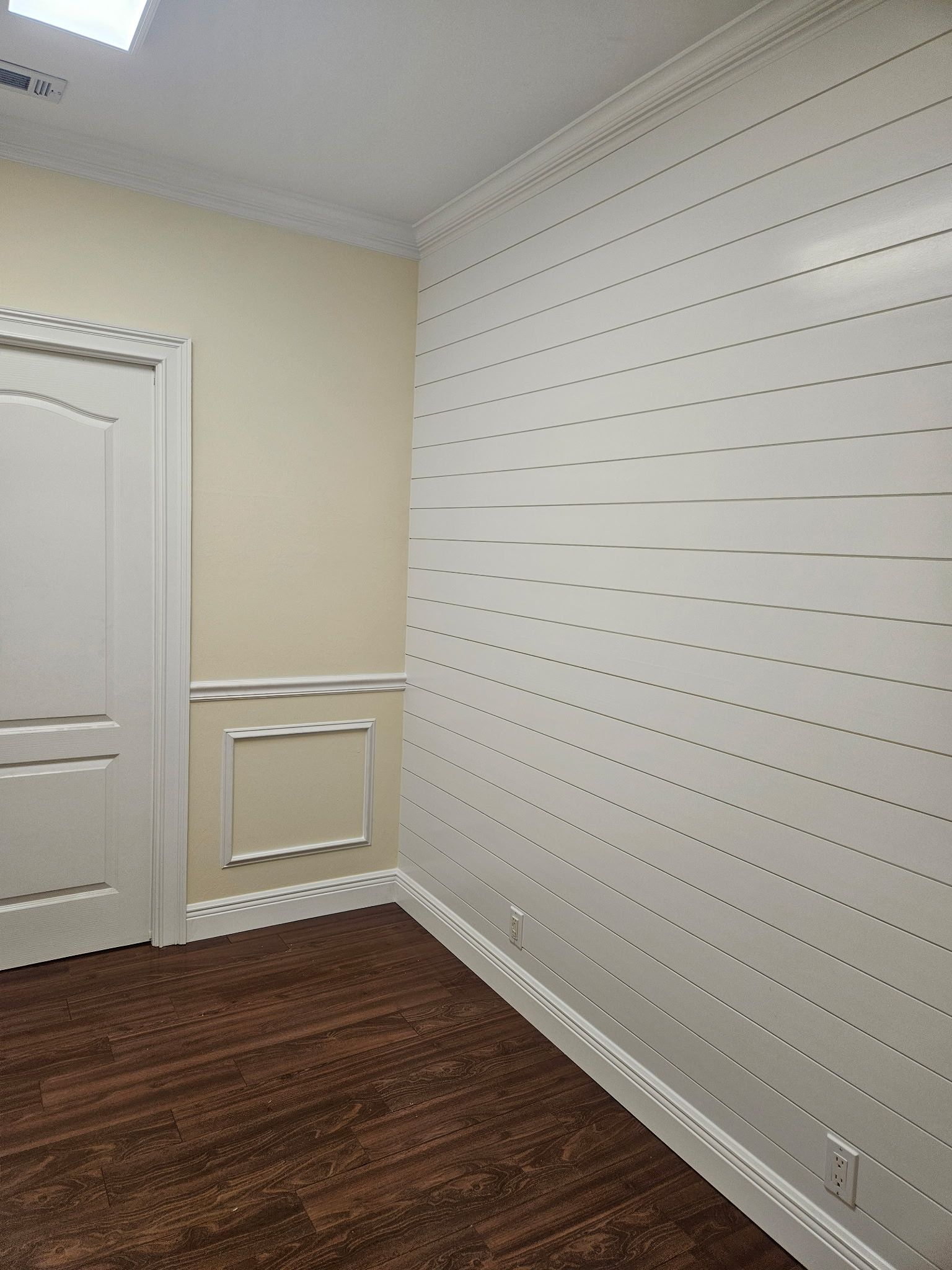 Corner of a room with white paneling and a wood-toned floor. A door, cream walls, and decorative molding are also visible.