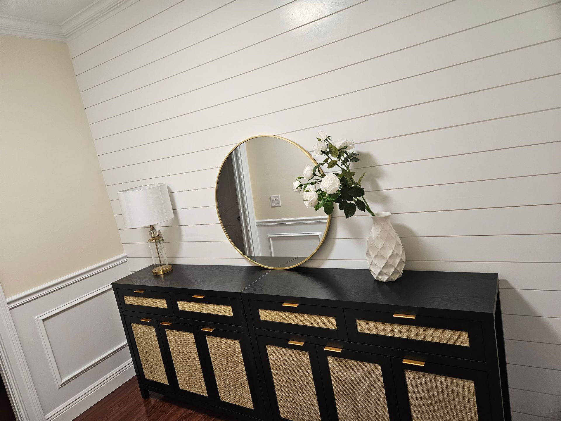 Black and gold sideboard with round mirror and white flower arrangement against a shiplap wall.