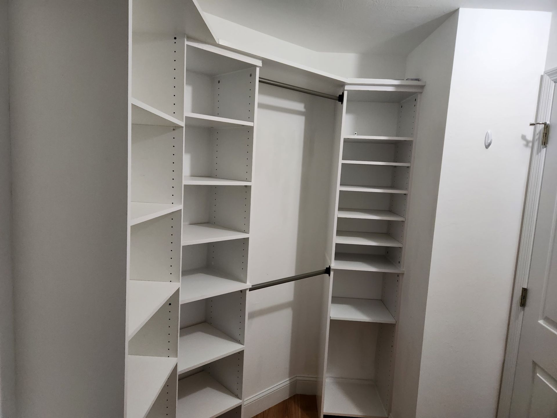 White built-in closet with shelves and a hanging rod. Shelves arranged in an L-shape. A door is visible on the right.