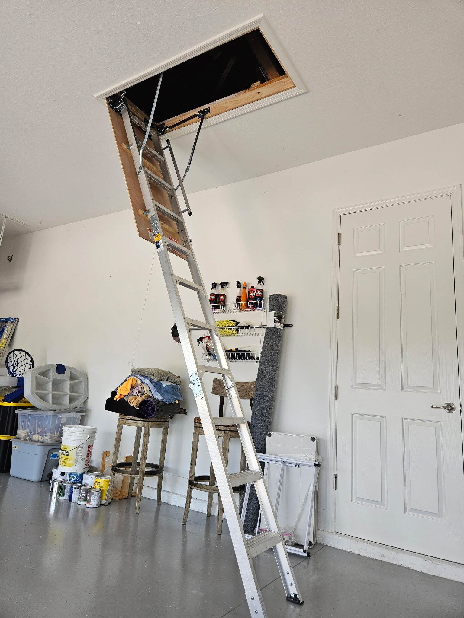 Attic access ladder extended from an open ceiling hatch in a garage with a white door and various items stored 