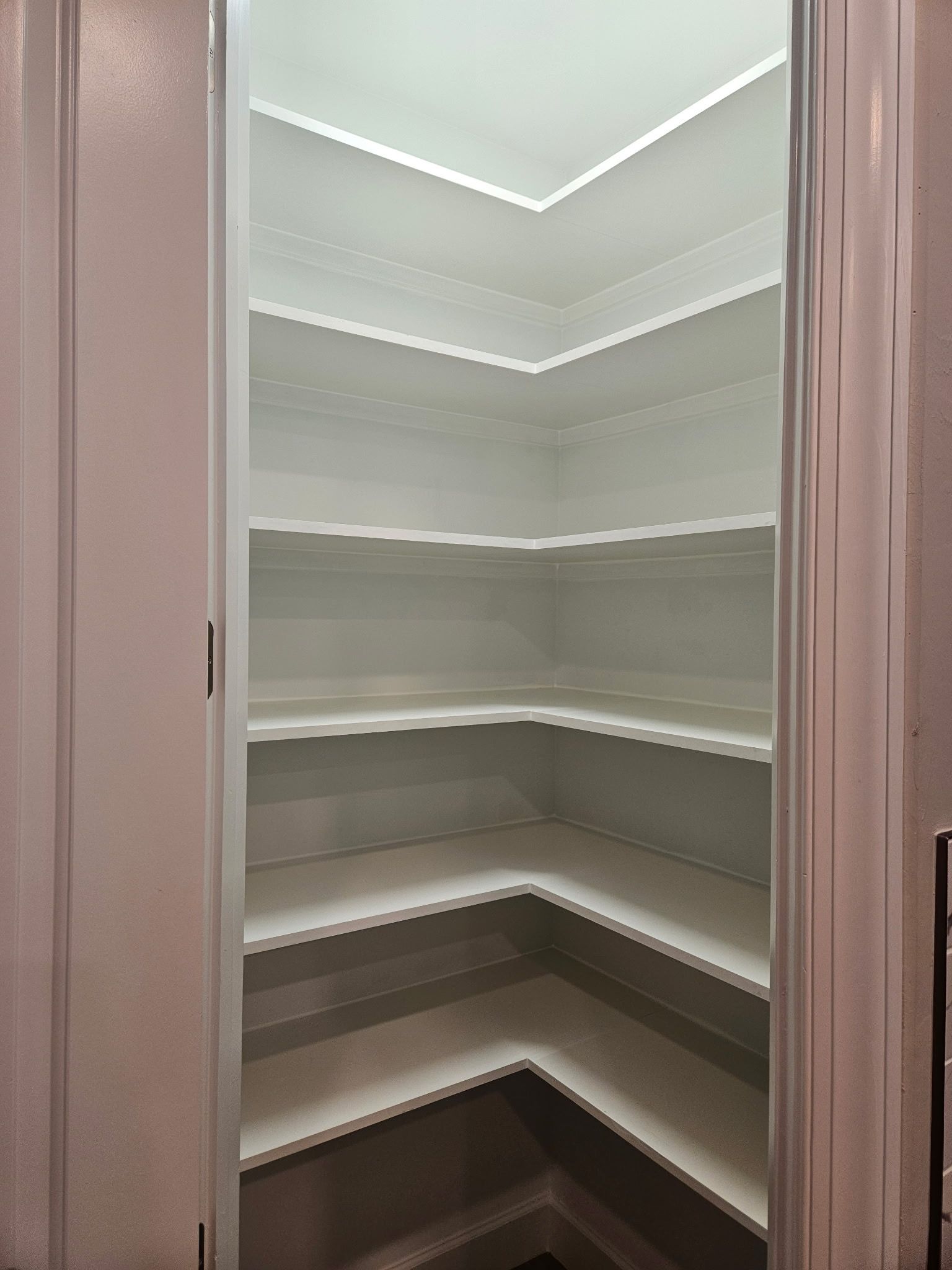 A white, empty pantry with several shelves, viewed from the doorway. The shelves are arranged in a corner formation.