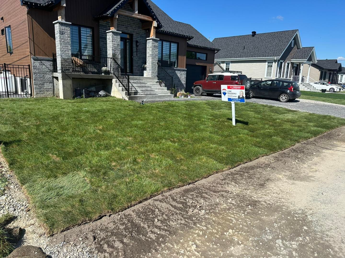 Pelouse fraîchement gazonnée devant une maison avec un panneau « à vendre » ; allée de gravier et rue visibles.