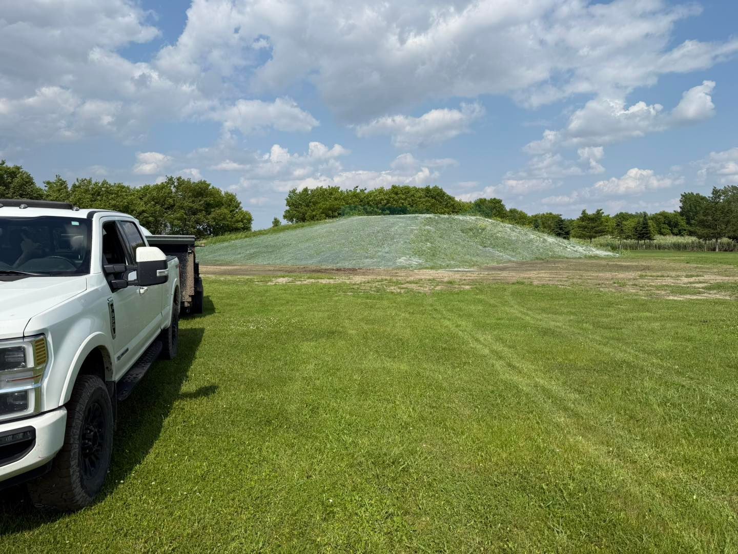 Camionnette blanche garée sur l'herbe verte, près d'un gros tas recouvert d'un filet vert sous un ciel partiellement nuageux.