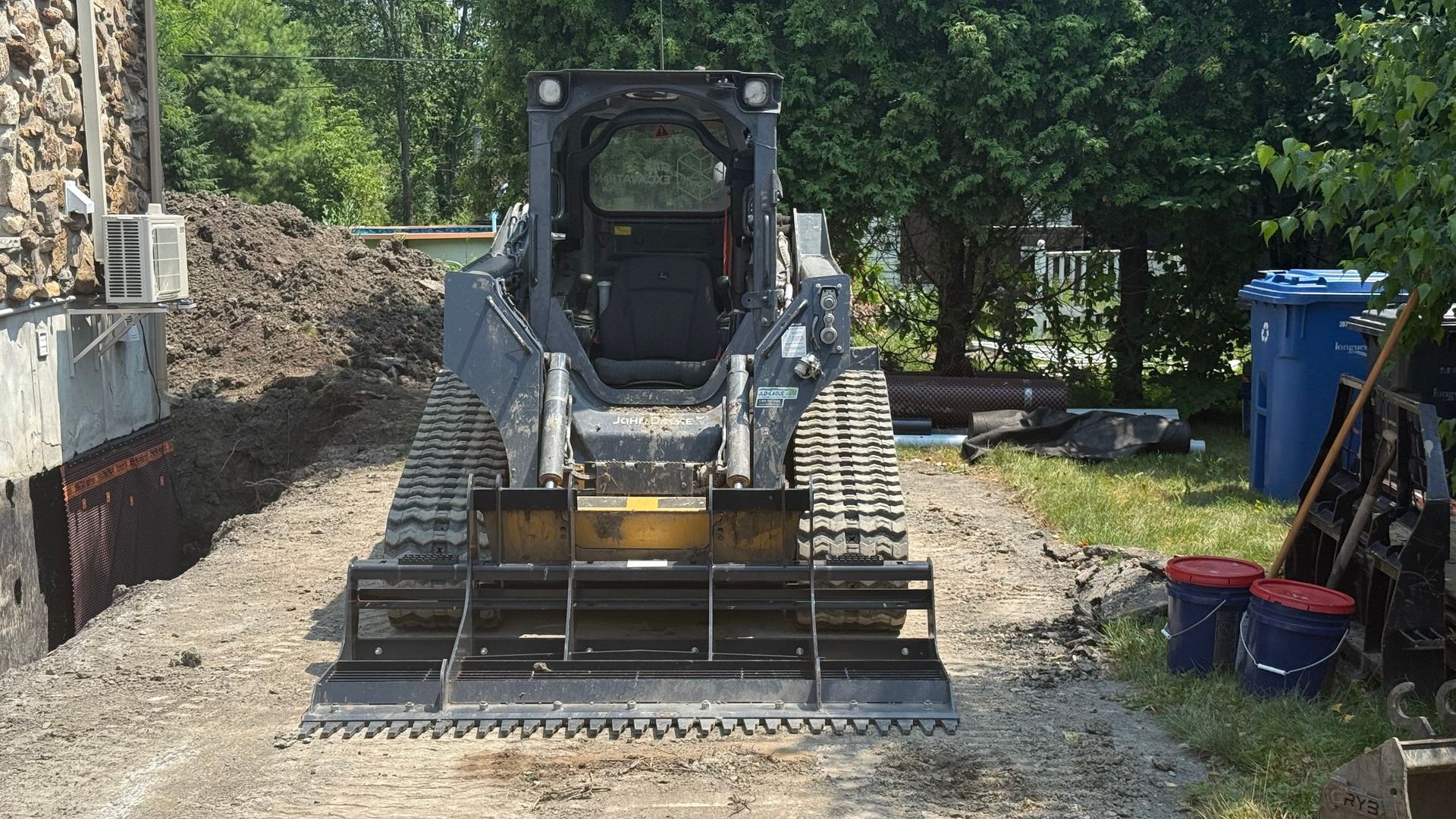Une chargeuse compacte grise à chenilles est garée sur du gravier près des fondations d'un bâtiment et d'un tas de terre.