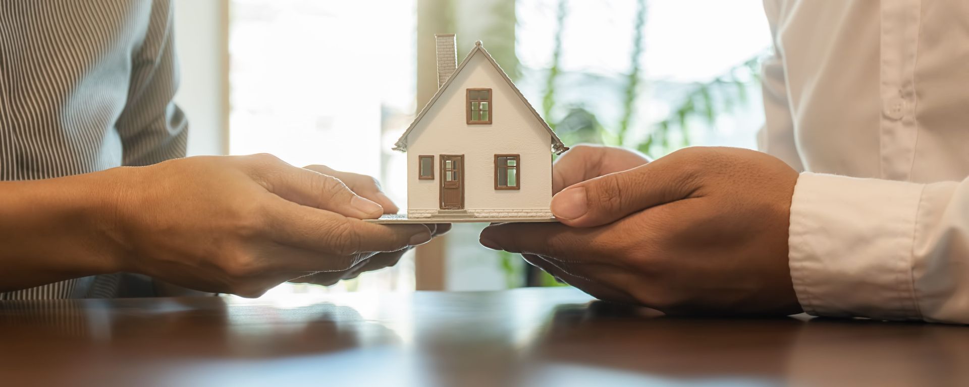 Two sets of hands hold a small model house between them on a table, symbolizing a real estate or property transaction.