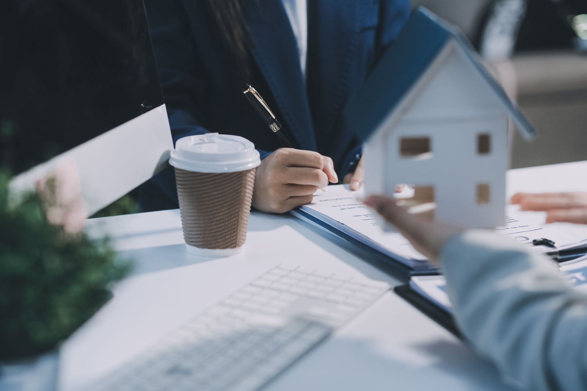 Hands with miniature house, pen, and documents at a desk; business meeting.