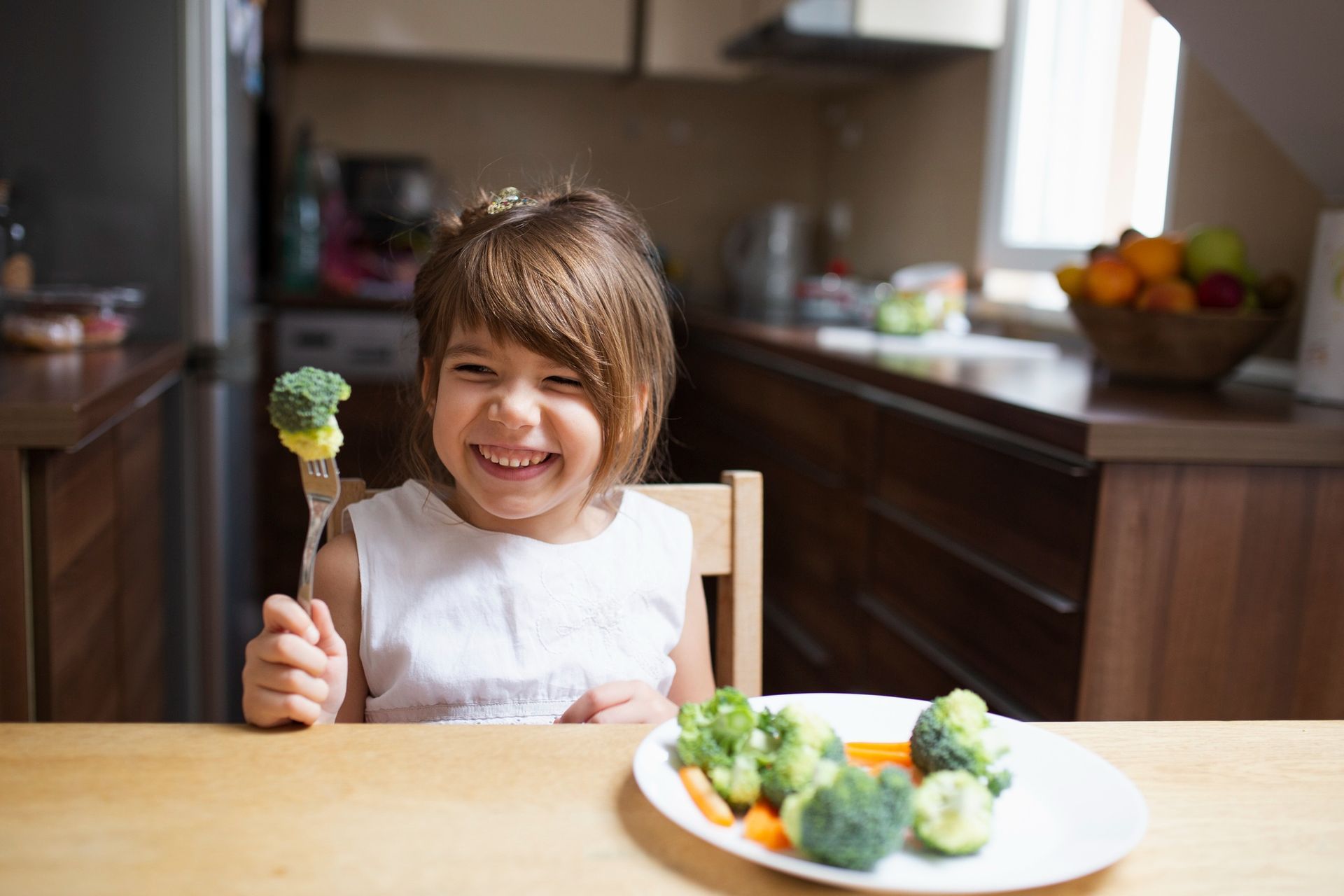 Smiling young girl sitting at a table, happily holding a fork with broccoli and a plate of fresh vegetables in front of her, representing pediatric nutrition and healthy eating habits.