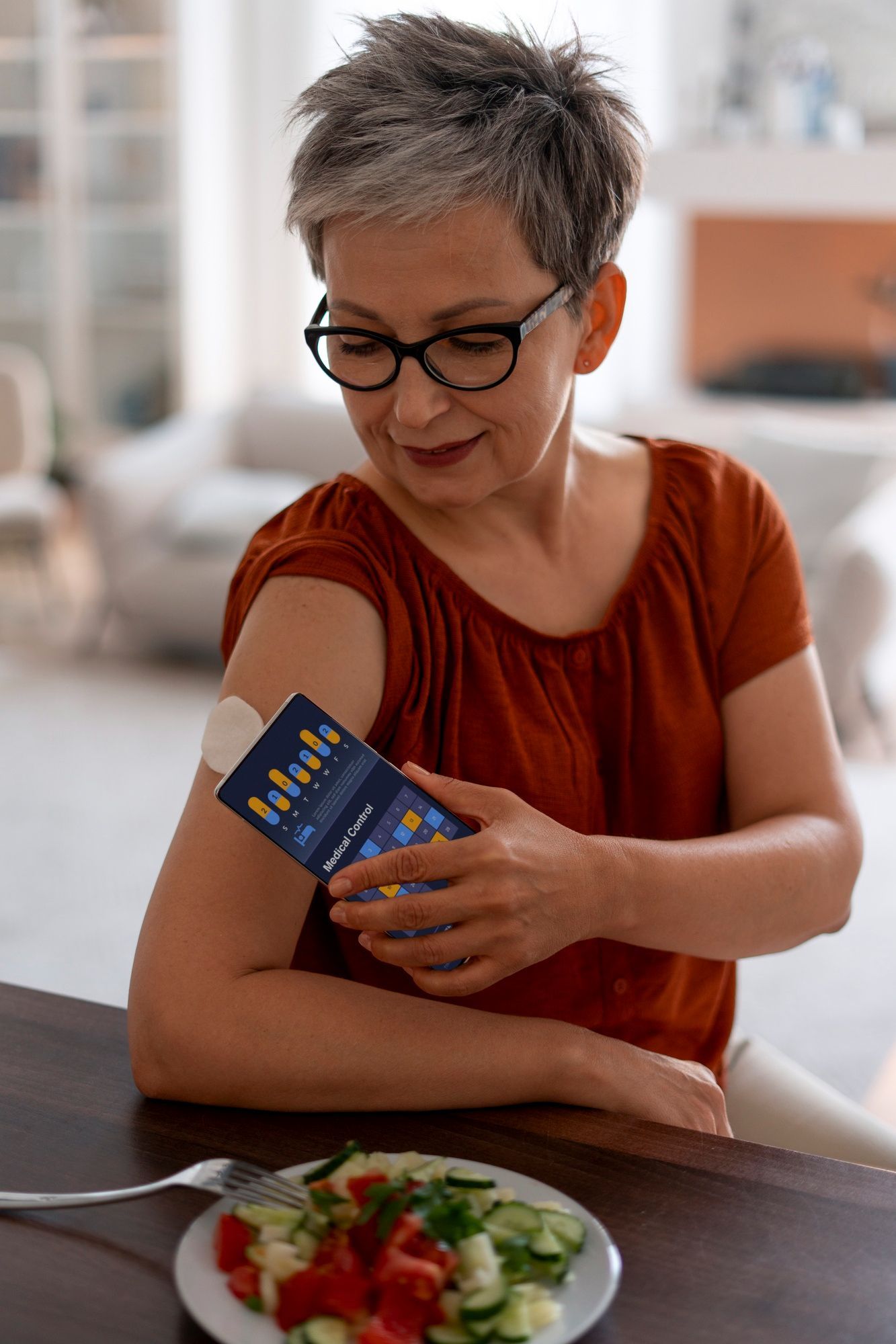Older woman checking her blood sugar with a digital glucose monitor on her arm while sitting at a table with a healthy vegetable salad, representing diabetes management and balanced nutrition.