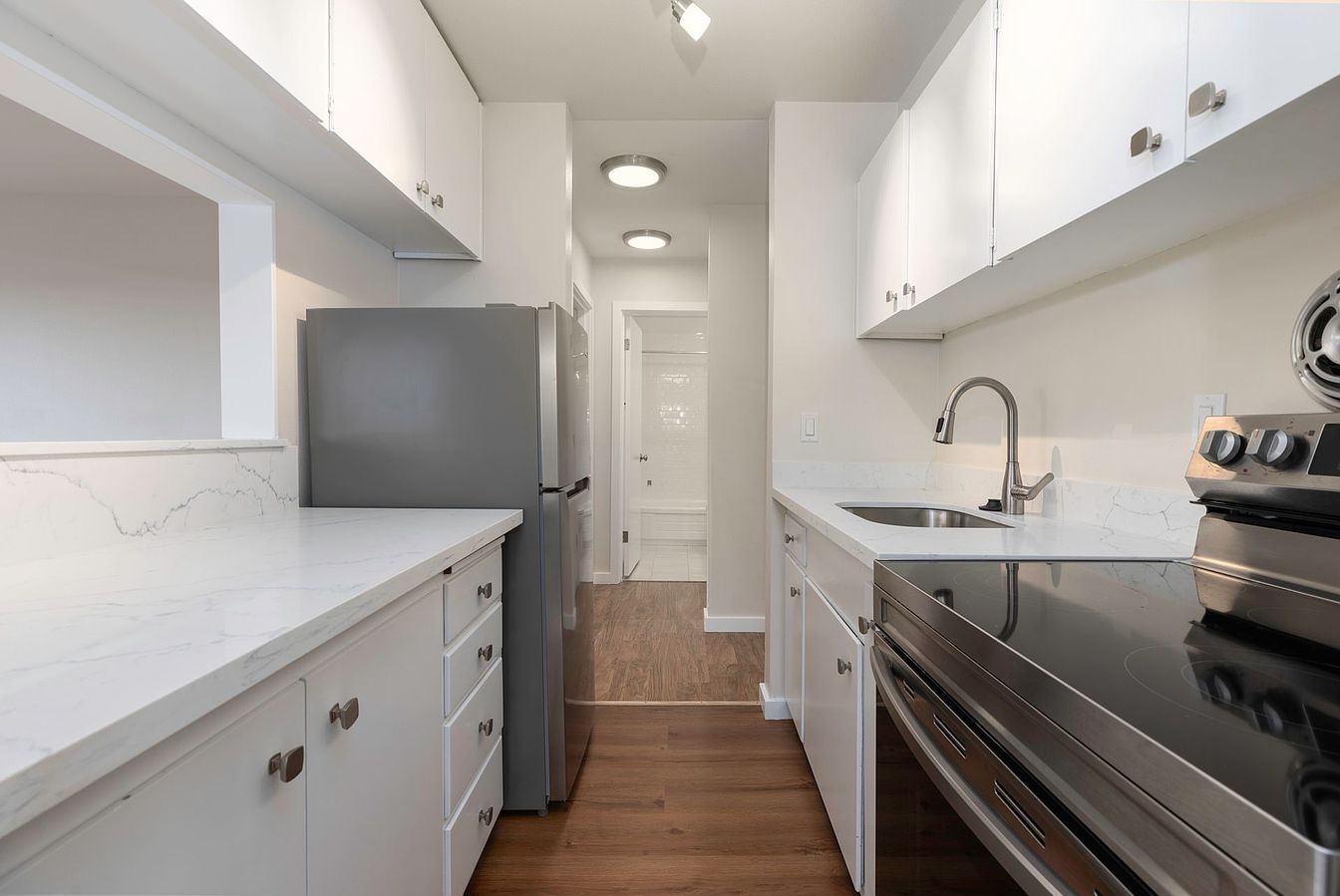 Narrow kitchen with white cabinets, stainless steel appliances, and a view of a hallway.