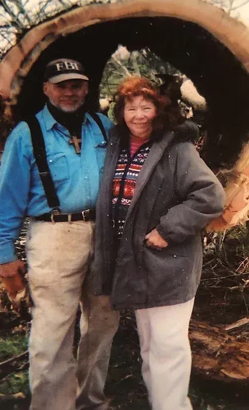 Man and woman pose in front of a large tree trunk. The man wears a hat, blue shirt, suspenders and the woman a grey jacket.
