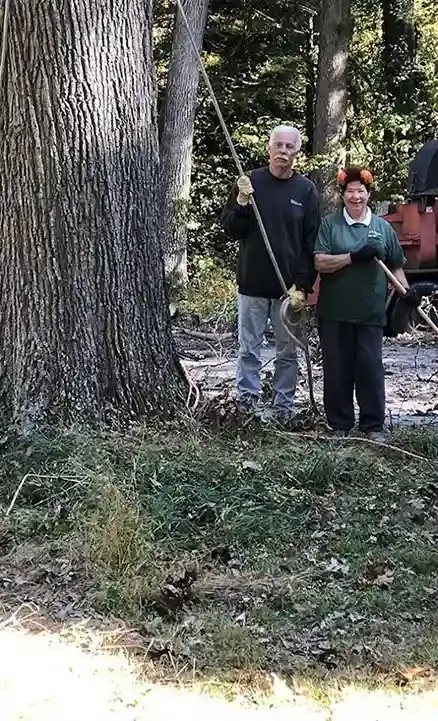 Two people stand near a tree, man holding a rope, woman with a tool; outdoors, green and brown tones.