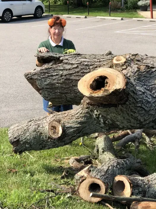 Woman stands next to large cut tree trunk in a parking lot, wearing orange earmuffs.