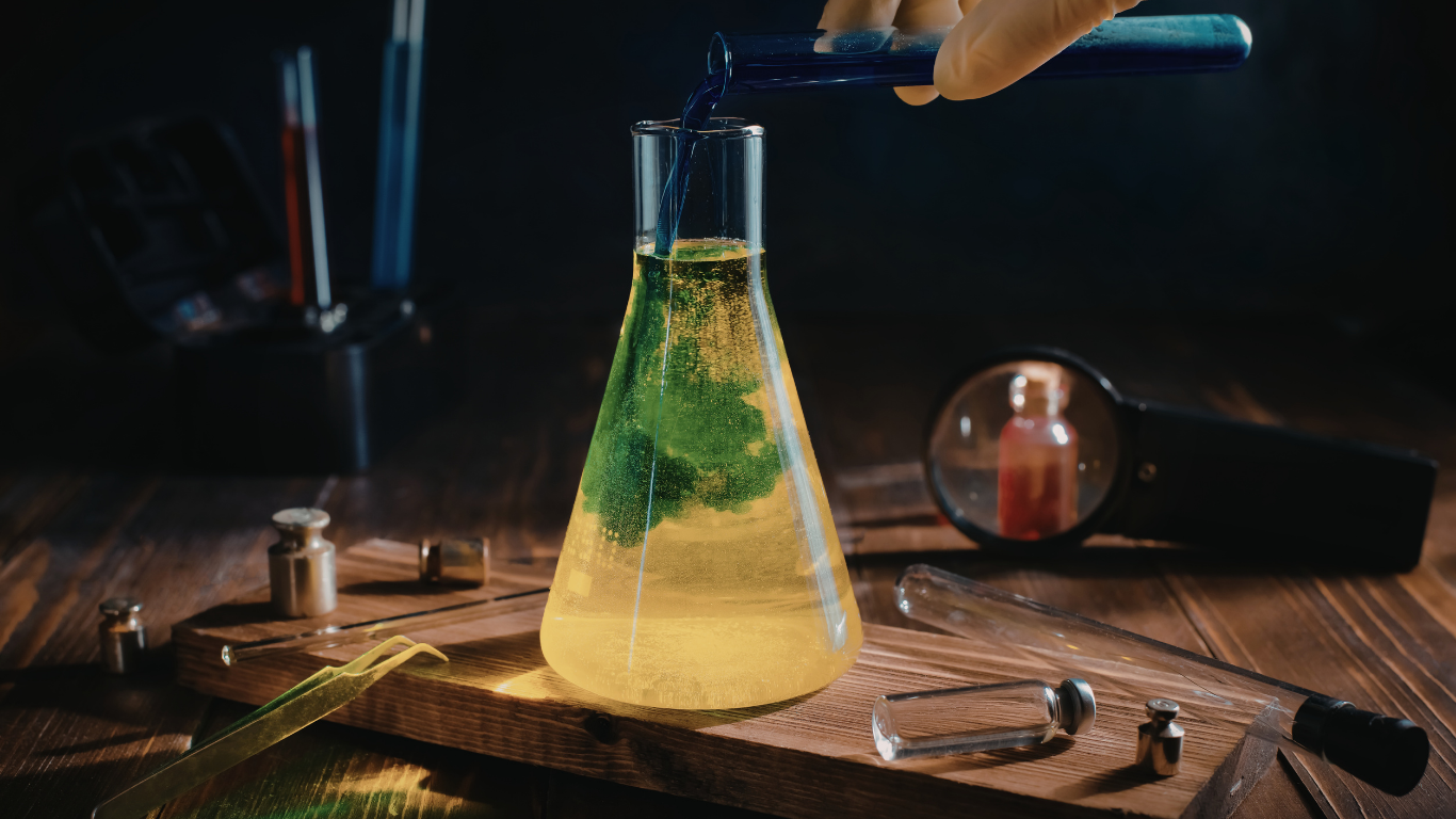 Person pouring blue liquid into a flask with yellow liquid and green substance, on wooden surface.