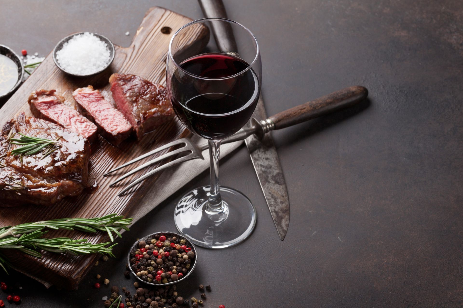 Sliced steak with rosemary, peppercorns, and a glass of red wine on a dark rustic table.