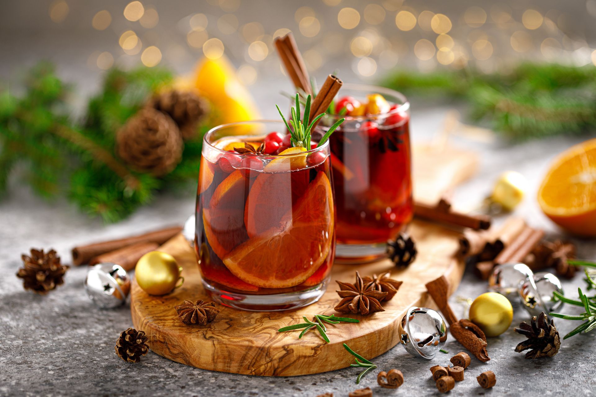 Two glasses of mulled wine with cinnamon sticks, orange slices, and cranberries on a wooden board with holiday decorations.