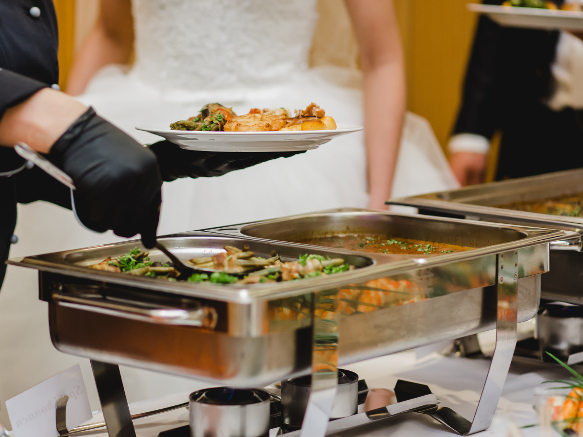 Caterer serving food from a buffet; bride in white dress in background.