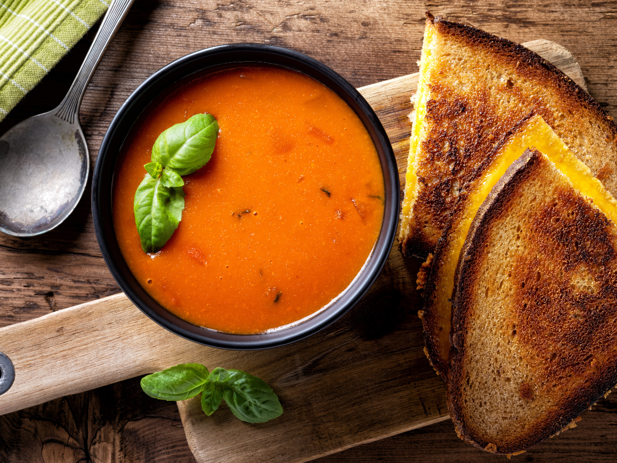 Tomato soup in a black bowl with basil, next to grilled cheese sandwich on wooden board.