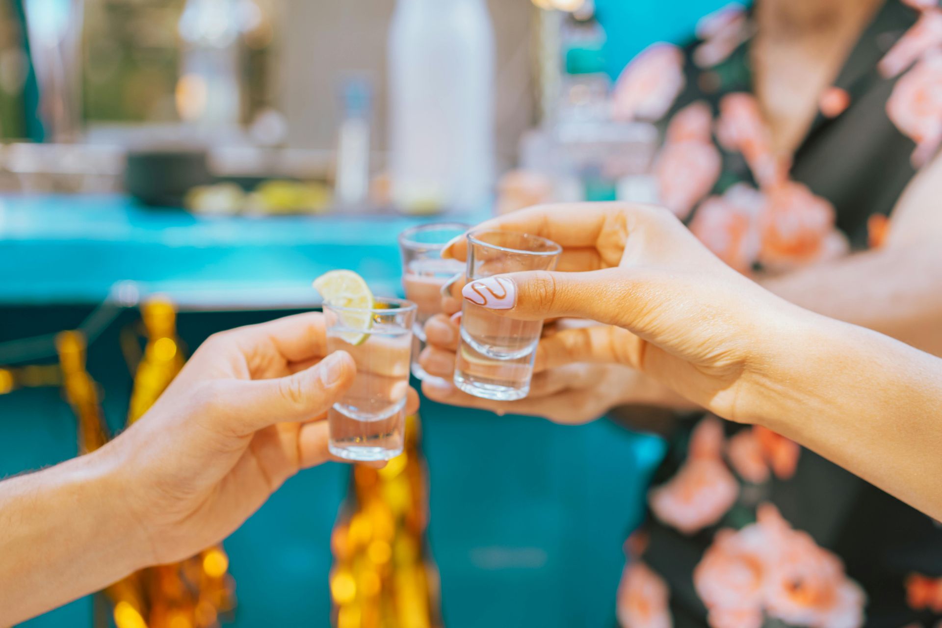 Hands toasting tequila shots, one with a lime wedge, against a blurred outdoor background.