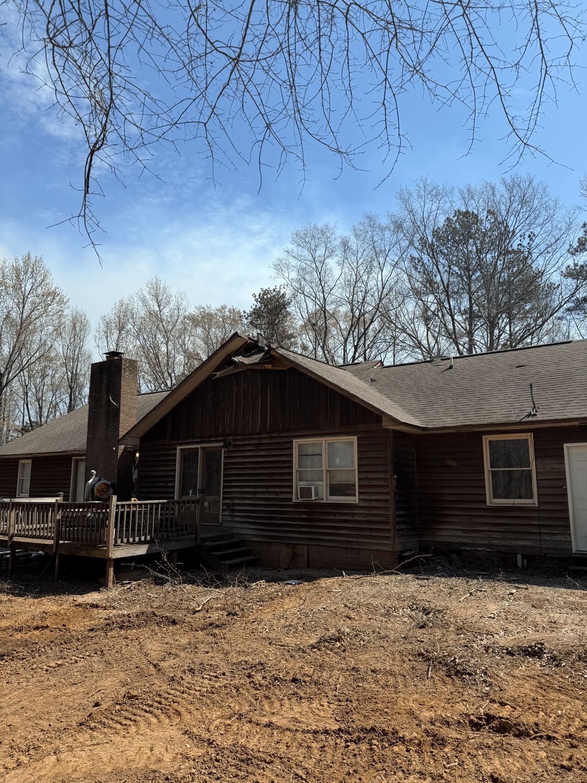 Log cabin with chimney, windows, and a deck against a blue sky, with trees.