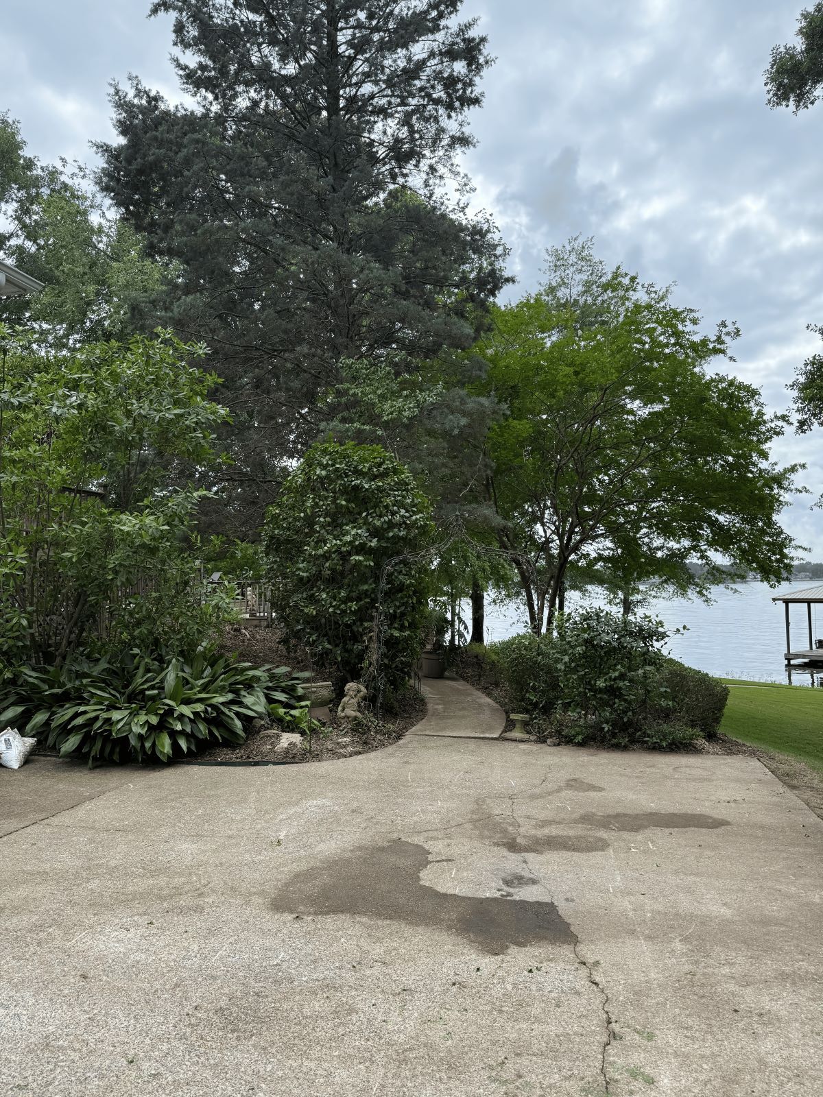 Concrete driveway leads to a lush, green waterfront with trees under a cloudy sky.
