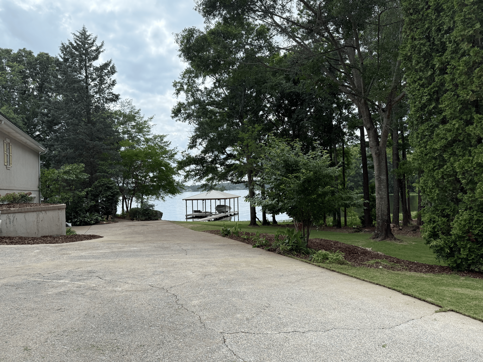 Concrete driveway leads to a lake with a boat dock, surrounded by trees under a cloudy sky.