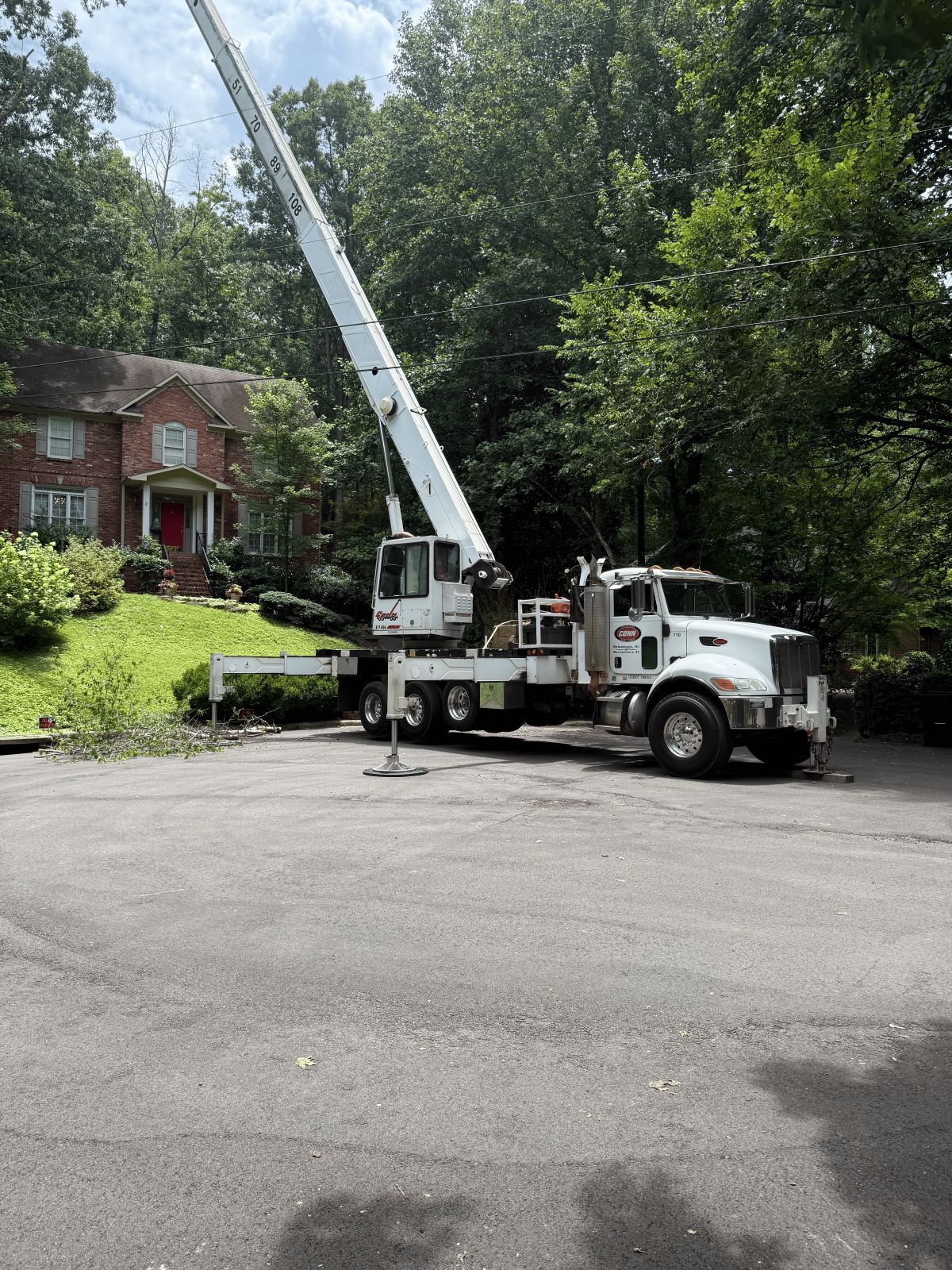 White tree service truck with crane extended in front of a two-story brick house on a hill.