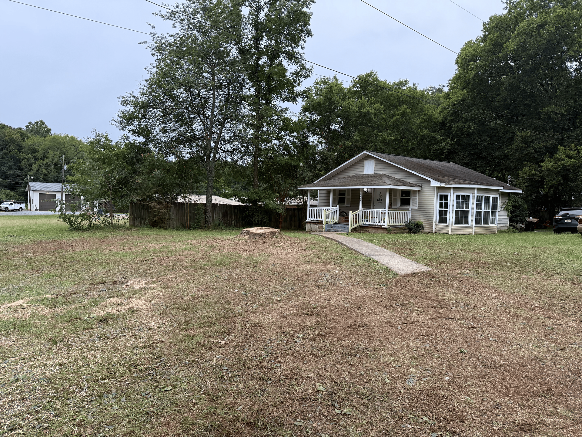 A small, weathered house with a porch and a pathway on a grassy lot. Trees and overcast sky visible.