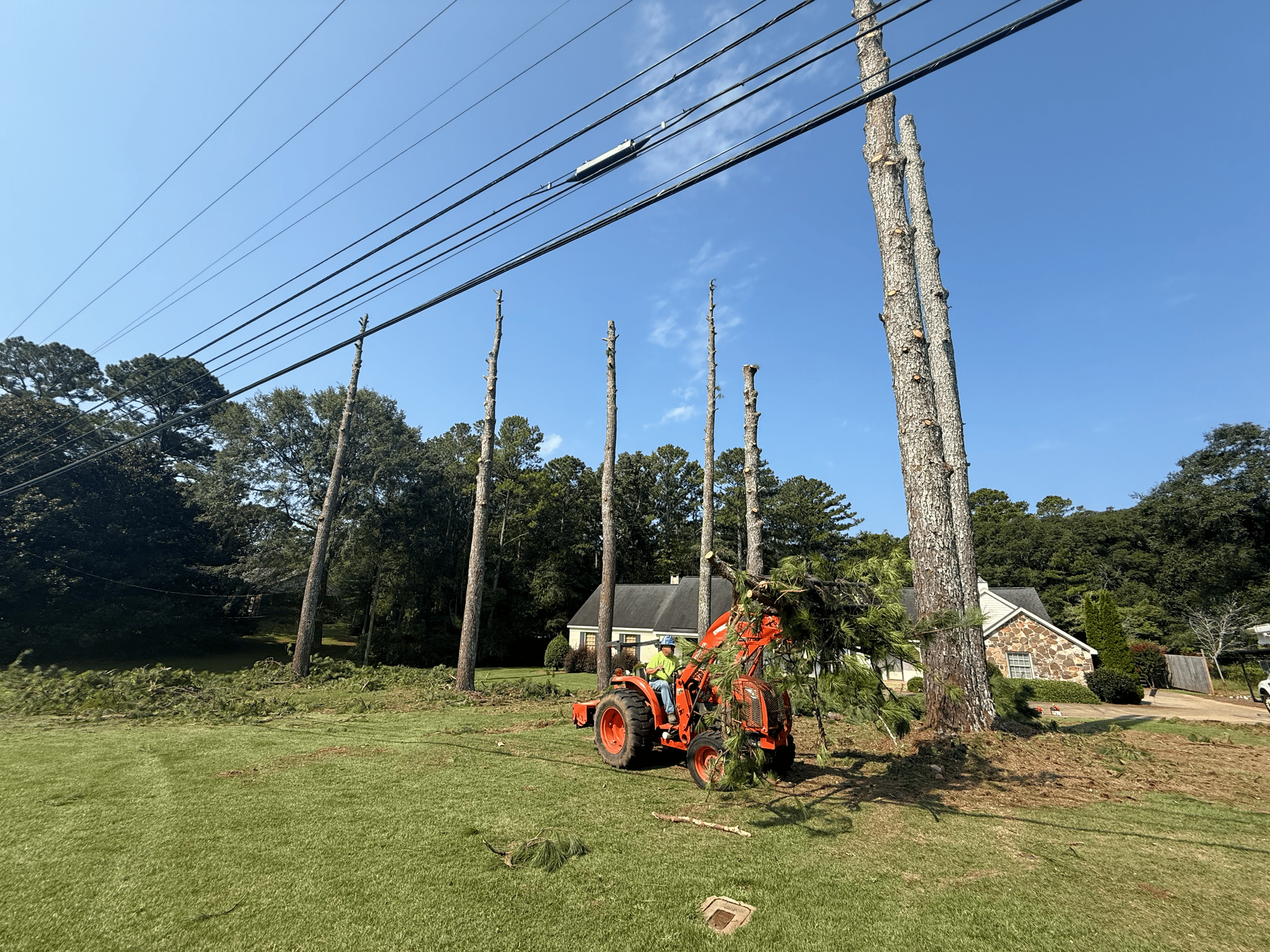 A tractor with a worker clearing brush near power poles and power lines under a blue sky.